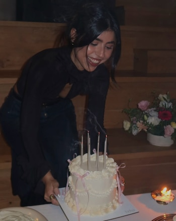 Cami Mayan, con el pelo oscuro recogido y blusa negra, sonríe ampliamente mientras sostiene un pastel blanco con velas apagadas y humo en una mesa