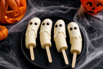Cuatro trozos de plátano en palitos de madera cubiertos con chocolate blanco y ojos negros sobre un plato, con calabazas y telarañas.