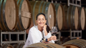 Mujer sonriente con cabello oscuro recogido, vestida de blanco, sostiene una copa de vino tinto. Se apoya en una barrica de roble con más barricas al fondo