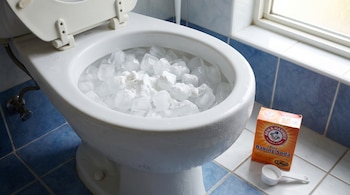 Un inodoro blanco lleno de cubos de hielo y polvo blanco, probablemente bicarbonato. Una caja de bicarbonato y una cuchara medidora están en el suelo.