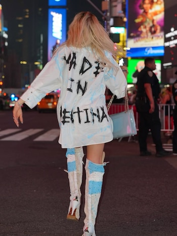 Mujer de espaldas con cabello rubio en calle de ciudad de noche. Viste camisa blanca con texto negro 'MADE IN CHINA' y botas blancas con azul. Lleva cartera azul claro