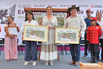 Dos mujeres y un hombre sostienen billetes de lotería enmarcados frente a una pancarta oficial, con una niña en uniforme rojo y otras personas en un escenario