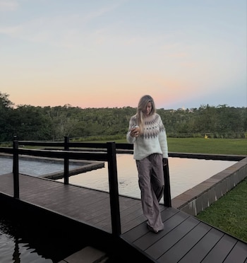 Mujer rubia camina por un puente de madera oscuro sobre agua, sosteniendo un mate. Viste suéter claro con patrón y pantalones oscuros, con vegetación y cielo rosado al fondo