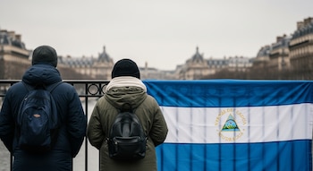 Vista trasera de dos personas con abrigos oscuros y mochilas, mirando una bandera de Nicaragua colgada de una reja en París, con edificios europeos borrosos al fondo.