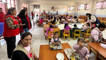 Interior de un comedor con niños sentados en mesas de madera y sillas pequeñas. Adultos supervisan a los niños. Algunos adultos usan chalecos rojos