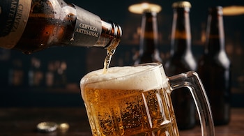 Una botella de cerveza artesanal vierte líquido dorado en una jarra de vidrio junto a otras botellas en el fondo.