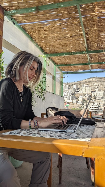 Una mujer teclea en una computadora portátil sobre una mesa de madera en una terraza con techo de paja y vista a edificios sobre una colina