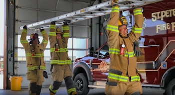 Tres bomberos equipados sostienen una escalera metálica sobre sus cabezas frente a un vehículo rojo de emergencia
