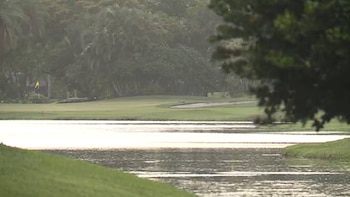 Vista panorámica de un campo de golf con un lago en primer plano, césped verde, un búnker de arena y árboles frondosos al fondo bajo un cielo claro