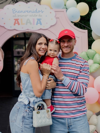 Alexis Mac Allister, Ailén Cova y una niña posan sonrientes frente a un arco decorado con globos y un cartel que dice Alaia