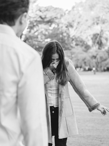 Una mujer de cabello largo, vestida con un abrigo forrado y pantalón oscuro, se cubre la boca con la mano en un gesto de sorpresa o emoción, al aire libre. Un hombre aparece de espaldas en primer plano, desenfocado