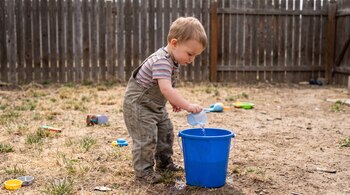 Niño pequeño con mono y camiseta a rayas, vertiendo agua de una taza a una cubeta azul en un patio de tierra y pasto. Valla de madera al fondo.