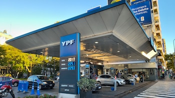 Vista frontal de una estación de servicio YPF con cartel de precios, surtidores, varios coches y una moto. Edificios y árboles al fondo bajo cielo azul
