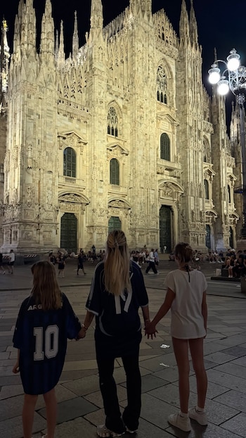 Mujer rubia y dos niñas de espalda, tomadas de la mano, miran una fachada de catedral gótica iluminada. Noche, gente y farola en la plaza