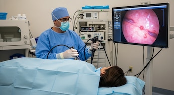 Un médico con mascarilla y guantes realiza una colonoscopia a un paciente acostado, mientras un monitor muestra el interior del colon con pólipos.