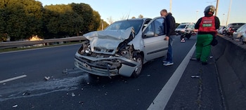 Un vehículo plateado gravemente dañado en la parte delantera está en una carretera, con un hombre de pie junto a él y un médico del SAME de espaldas
