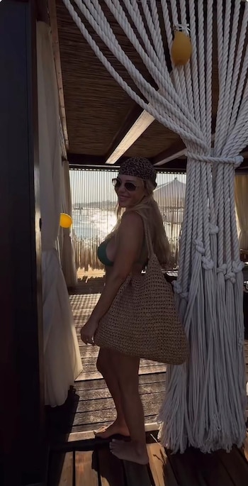 Mujer rubia sonriendo con gafas de sol, pañuelo, top de bikini verde, bolso de paja, descalza en piso de madera. Al fondo, mar y cielo