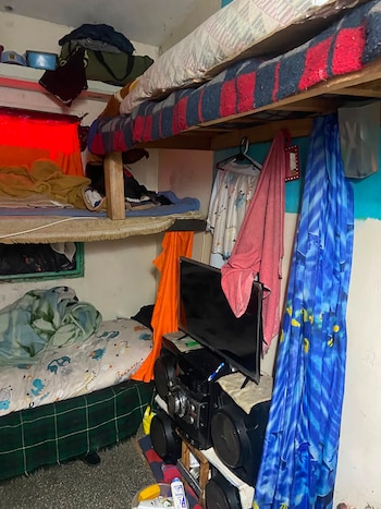 Habitación interior con literas, colchones, sábanas y mantas. Textiles colgados, ropa, toallas, un televisor y un equipo de sonido. Objetos varios