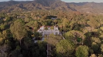 Vista aérea de una gran casa blanca de estilo clásico con tejado rojo, rodeada de exuberante vegetación y palmeras, con montañas al fondo