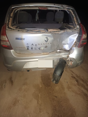 Vista trasera de un automóvil Renault Sandero plateado severamente dañado con el vidrio trasero roto, maletero abollado y parachoques desprendido, en un camino de tierra oscuro