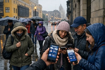 Personas con ropa abrigadora y paraguas caminan bajo la lluvia en una calle mojada de Lima. Algunos revisan la aplicación del Senamhi en sus teléfonos.