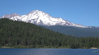Montaña nevada en la distancia, densos árboles verdes en una ladera, un lago azul en primer plano y cielo despejado. Un bote con dos personas