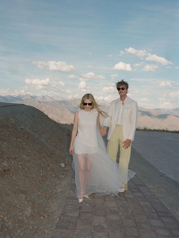 Una mujer y un hombre, Emma Roberts y Cody John, caminan de la mano por un sendero adoquinado al lado de un terraplén de tierra con montañas al fondo y un cielo con nubes