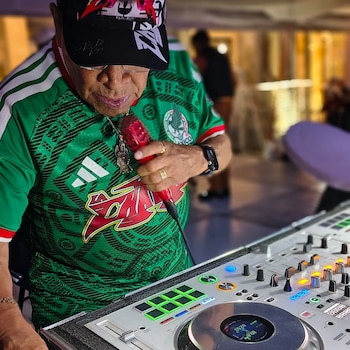 Un hombre con camiseta verde, gorra y gafas sostiene un micrófono rojo frente a una consola de mezclas