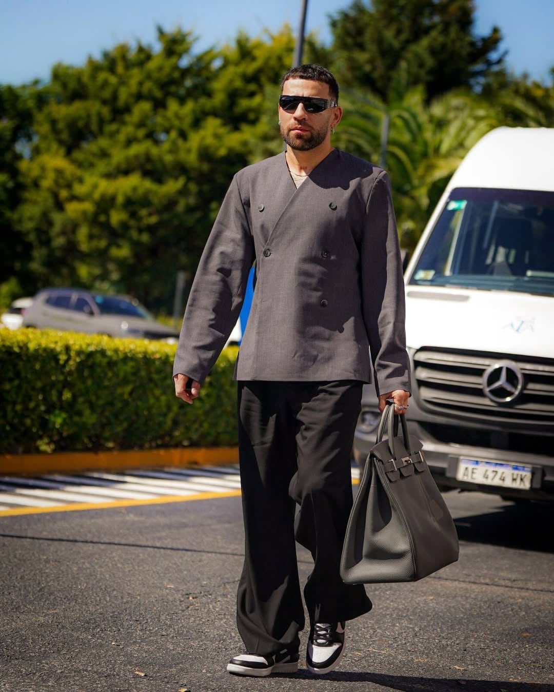 Nicolás Otamendi camina con un atuendo moderno de chaqueta gris, pantalones anchos negros y gafas de sol, llevando un bolso a juego