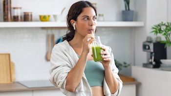 Mujer con auriculares y ropa deportiva que bebe un batido verde con una pajita desde una botella de vidrio en una cocina