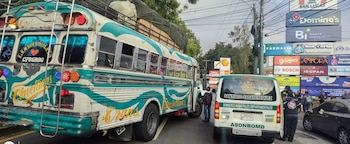 Autobús blanco con líneas turquesas y carga en el techo junto a una furgoneta blanca de Bomberos Municipales. Varias personas, carteles comerciales y un automóvil