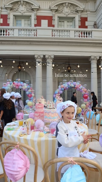 Una niña con gorro de chef y delantal sonríe frente a una mesa con pasteles decorados, farolillos y arcos de globos en un patio