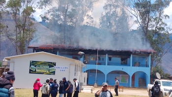 Un edificio escolar azul emite humo desde el techo mientras un grupo de personas permanece en el patio junto a una camioneta