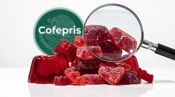 Varios dulces y gelatinas rojas apilados sobre una superficie blanca, una lupa de laboratorio los magnifica y el logo de Cofepris verde se ve al fondo.
