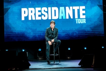 Dante Gebel, un hombre con cabello castaño oscuro y traje negro, sonríe mientras está sentado en un taburete alto en un escenario oscuro, con una pantalla azul brillante que dice "PRESIDENTE TOUR" detrás