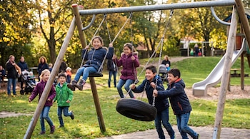 Un grupo de niños juega en un parque con columpios. Dos niñas se columpian, dos niños empujan un columpio de neumático. Otros niños y adultos en el fondo, árboles con hojas amarillas.