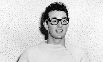 Retrato en blanco y negro de Buddy Holly con gafas de montura negra y suéter claro frente a una pared perforada