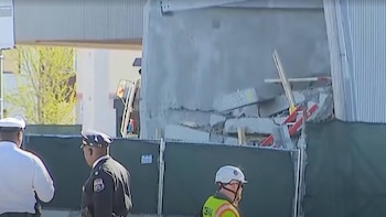 Dos oficiales de policía y un trabajador de construcción se encuentran cerca de una valla verde frente a una estructura gris parcialmente colapsada con escombros y elementos metálicos