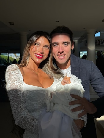 Una mujer con vestido blanco de encaje y un hombre con saco oscuro sonríen junto a un bebé envuelto en una manta blanca