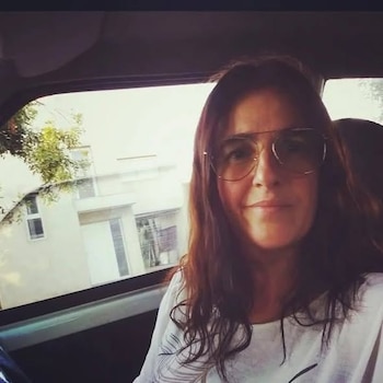 Una mujer de cabello castaño largo y gafas de sol, sonriendo levemente, tomada dentro de un coche. Un edificio blanco es visible a través de la ventana
