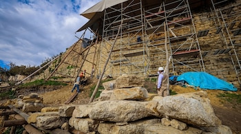 Trabajadores con cascos de seguridad junto a un gran muro de piedra rodeado por andamios metálicos, lonas y rocas en el suelo.