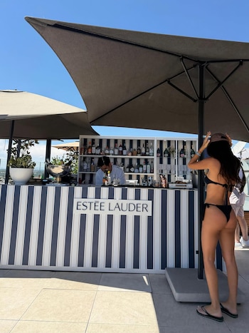 Mujer de espaldas en bikini negro con gorra y ojotas frente a una barra de bar con el logo Estee Lauder y botellas. Hay un camarero y sombrillas