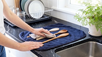 Manos de una persona que seca cucharas de madera sobre un tapete azul en una mesada de cocina junto al fregadero.