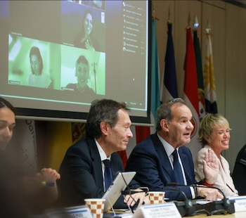 Tres panelistas presenciales, dos hombres y una mujer, sentados en una mesa con micrófonos, frente a una pantalla con cuatro participantes virtuales