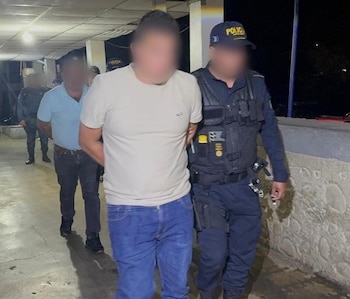 Un hombre esposado es escoltado por un policía junto a una camioneta oscura. Al fondo, un SUV rojo y un coche gris en la oscuridad de la noche