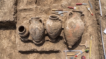 Cuatro vasijas de cerámica enterradas en tierra, rodeadas de herramientas de excavación como palas, brochas, reglas métricas y cintas de medir.