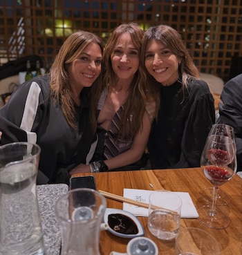 Tres mujeres sonrientes, incluyendo Verónica Lozano, posan en una mesa de restaurante con bebidas, palillos y salsa. Fondo con enrejado de madera