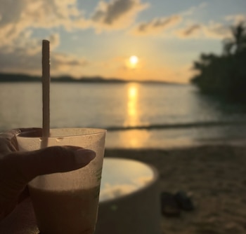 Una mano sostiene un vaso de plástico con pajilla frente al mar durante la puesta de sol en una playa con vegetación