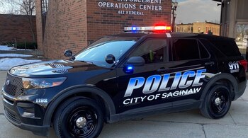 Un SUV policial negro del Departamento de Policía de Saginaw, con luces rojas y azules encendidas, estacionado frente a un edificio de ladrillos con nieve en el suelo