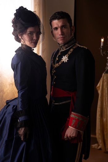 Hombre y mujer posan. Él viste uniforme azul con detalles dorados, banda roja y medalla. Ella lleva vestido azul oscuro, sombrero negro con velo y guantes de encaje